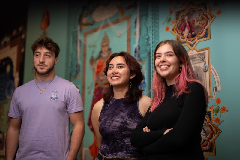 Three students smile in front of a colourful backdrop.